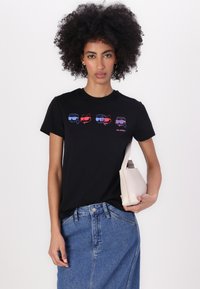 Black cotton t-shirt with short sleeves featuring colorful cartoon faces wearing sunglasses, paired with a denim skirt and small beige bag.