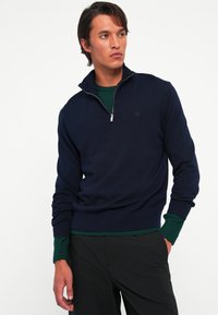 Mann iført en marineblå quarter-zip genser over en grønn langermet skjorte og svarte bukser, står med den ene hånden i lommen og ser til siden.