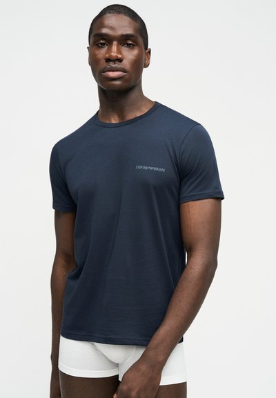 Νεαρός μαύρος άνδρας που φοράει μπλε σκούρο t-shirt Emporio Armani και λευκά ανδρικά εσώρουχα, στέκεται μπροστά σε ένα απλό φωτεινό φόντο.