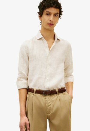 Homme aux cheveux bouclés foncés portant une chemise beige rayée à manches longues, un pantalon kaki plissé et une ceinture en cuir marron, debout les mains dans les poches.