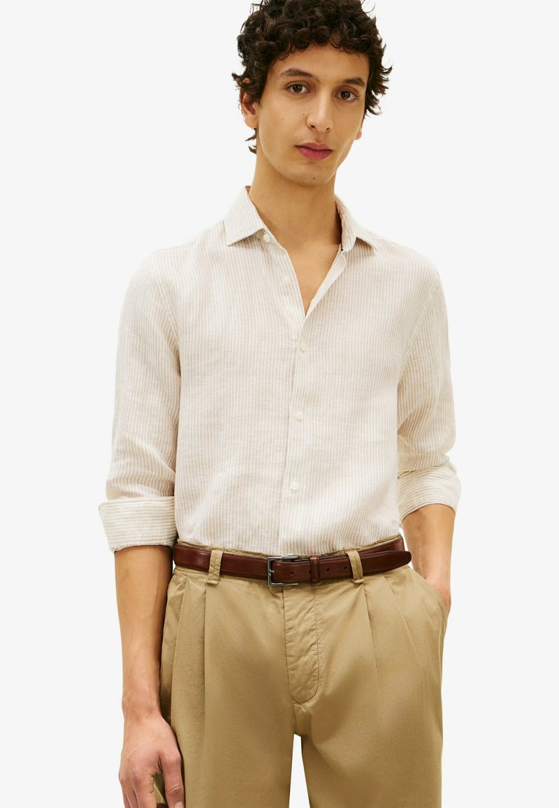 Homme aux cheveux bouclés foncés portant une chemise beige rayée à manches longues, un pantalon kaki plissé et une ceinture en cuir marron, debout les mains dans les poches.