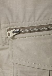 Béžová tkanina s mřížkovým vzorem, obsahující stříbrný zip, vrstvený design kapes a viditelné švy. Hladká, lehká struktura.