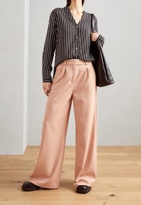 Černobílé pruhované tričko s výstřihem do V, s knoflíkovým detailem, v kombinaci se světle růžovými širokými kalhotami a černými botami, nese černou tašku.