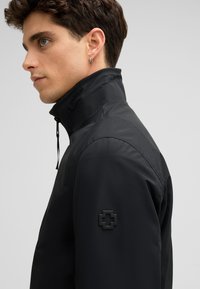 Schwarze Jacke mit hohem Kragen und Reißverschluss, aus glattem Stoff mit matter Textur. Enthält ein schwarzes Logo-Patch auf der Schulter.