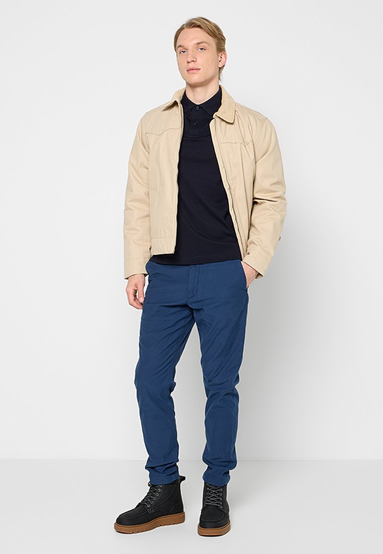 Veste légère beige portée sur un polo marine, associée à un pantalon bleu et des bottes à lacets noires avec une semelle brune, se tenant devant un fond uni.