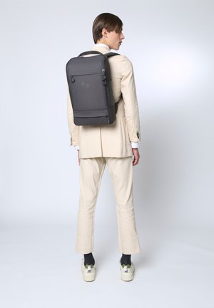 Schwarzer rechteckiger Rucksack mit zwei oberen Griffen, Reißverschluss und minimalistischen Design. Glattes Material; verfügt über zwei kreisförmige Akzente auf der Rückseite.