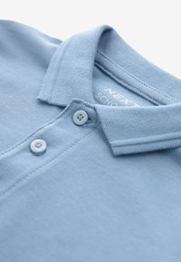 Polo bleu clair à manches courtes avec col plié et deux boutons sur la patte de boutonnage, montrant une partie du texte de l'étiquette intérieure.