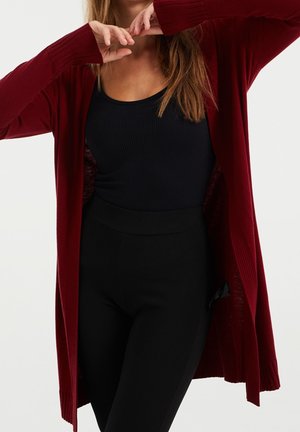Femme portant un long cardigan rouge par-dessus un haut noir sans manches et un pantalon noir taille haute, avec les mains près du visage.