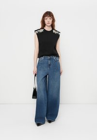 Svart ermeløs topp med dekorative detaljer på skuldrene, sammen med vide blå jeans og en liten svart håndveske. Svarte spisse sko fullfører antrekket.