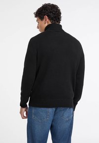 Maglione nero a maglia con colletto alto a coste e orlo a coste, abbinato a jeans in denim blu chiaro. La texture sembra morbida e accogliente.