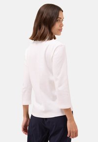 Maglietta bianca a maniche lunghe con scollatura rotonda e vestibilità rilassata. Presenta una texture liscia e opaca, mostrata di spalle con capelli di lunghezza spalle.