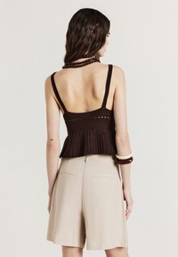 Donna con lunghi capelli castani, che indossa una canotta di maglia senza maniche marrone scuro, pantaloni pantaloni beige a vita alta e braccialetti spessi sul braccio destro.