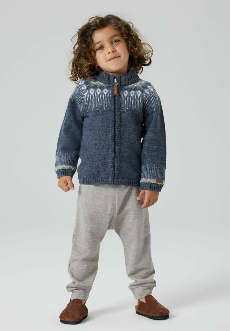 Kind met krullend haar draagt een blauw patroon met ritssluiting, lichte grijze broek en bruine instappers, staande tegen een effen achtergrond.