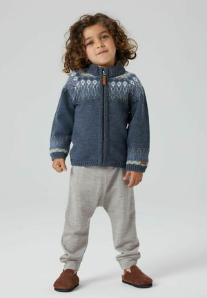 Kind met krullend haar draagt een blauw patroon met ritssluiting, lichte grijze broek en bruine instappers, staande tegen een effen achtergrond.