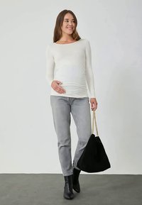 Mulher grávida com uma blusa de mangas longas branca, jeans cinzentos, botas pretas, segurando uma mala preta com corrente dourada, sorrindo e olhando para a direita.