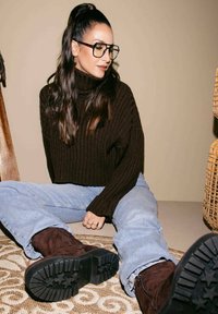 Maglione a collo alto a coste marrone, jeans azzurri chiari e stivali scamosciati marrone scuro. Il motivo texturizzato su un tappeto chiaro è visibile sotto.