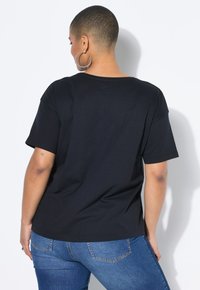 Persoon met kort haar die grote hoepeloorbellen draagt, een eenvoudig zwart T-shirt met korte mouwen en blauwe jeans, van zich afgekeerd tegen een witte achtergrond.