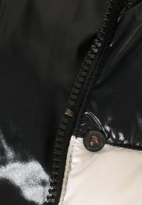 Gros plan sur une fermeture éclair noire et un bouton-pression noir sur un tissu brillant noir et blanc, probablement partie d'une veste ou d'un manteau.