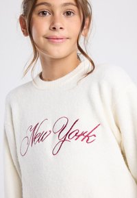 Pull crème à col rond, orné d'un texte brodé "New York" en rouge dans un style cursif. Tissu doux et texturé.