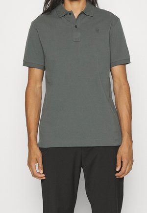 Mand iført en mørkegrå poloshirt med korte ærmer og to knapper, kombineret med sorte bukser, stående foran en ensfarvet baggrund.