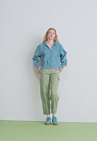 Modrá mikina na zips s predným vreckom, skombinovaná so svetlozelenými cargo nohavicami. Obsahuje modré tenisky s béžovou podrážkou, postavená proti jednoduchému pozadiu.