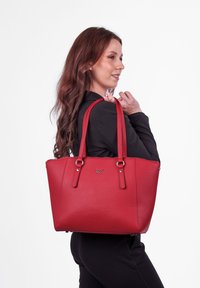 Červená kožená taška přes rameno s dvojitými ramenními popruhy, texturovaným povrchem a stříbrnými doplňky. Má zaoblené hrany a zipové uzavírání.