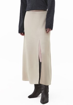 Béžová maxi sukně s předním rozparkem, vyrobená z měkké látky. Je zkombinována s přiléhavými černými botami s elegantním designem a špičatou špičkou.