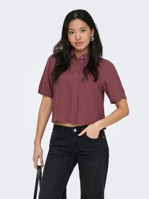 Femme aux longs cheveux foncés portant une chemise bordeaux courte à manches courtes boutonnée et un jean noir, tenant une lanière de sac noire.