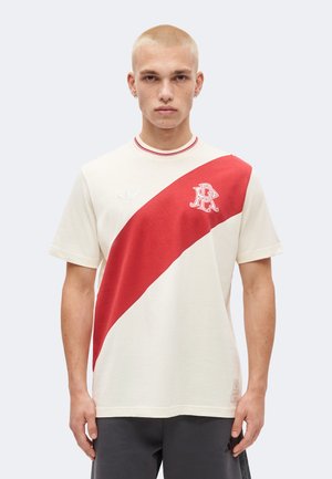 Jeune homme aux cheveux blonds courts portant un t-shirt blanc avec une large bande diagonale rouge et un logo sur la poitrine gauche, debout devant un fond uni.