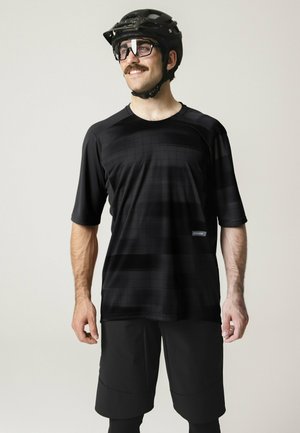 Mann trägt schwarzen Fahrradhelm, Brille, kurzärmliges schwarzes Trikot und schwarze Shorts, lächelt und schaut leicht zur Seite.