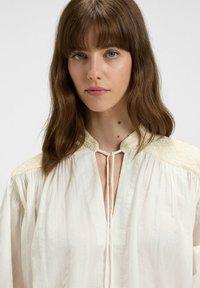 White blouse with intricate textured fabric, a keyhole neckline, and detailed embroidery on the shoulders. Loose, flowing design.