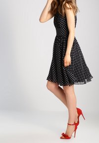 Schwarzes Polka-Dot-Kleid mit tailliertem Oberteil, schwingendem Rock und transparenter Saum. Kombiniert mit roten High-Heels, die eine Schleifenapplikation haben.