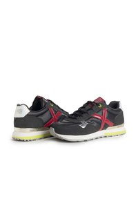 Zapatillas deportivas negras con acentos rojos y ante gris, que presentan una parte superior texturizada, suela de goma y un detalle amarillo en los cordones.