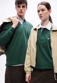 Δύο νέοι ενήλικες που φοράνε πράσινα ράγκμπι πουκάμισα Tommy Jeans και μπεζ μπουφάν, στέκονται μπροστά σε λευκό φόντο.