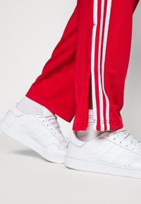 Röda träningsbyxor med vita sidostreck, kombinerat med vita sneakers. Sneakers har en texturerad yta och tre ränder på sidan.