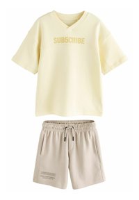 Chemise à manches courtes jaune clair avec le texte "SUB5CRIBE", associée à un short beige avec une taille élastique et des poches latérales.