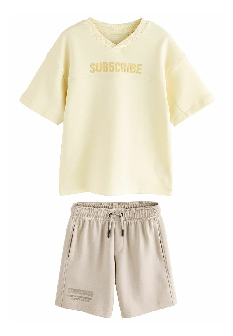 Chemise à manches courtes jaune clair avec le texte "SUB5CRIBE", associée à un short beige avec une taille élastique et des poches latérales.