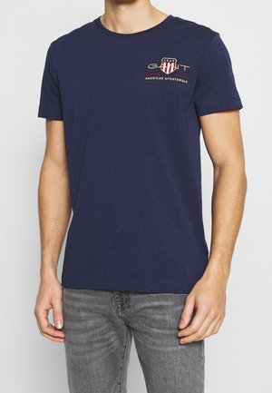 Mężczyzna noszący granatowy t-shirt GANT z logo i napisem „American Sportswear”, w połączeniu z szarymi dżinsami, stojący na białym tle.