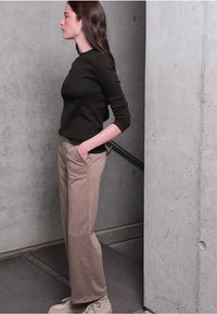 Haut à manches longues noir, pantalon beige à motifs et chaussures claires. Le mannequin se tient devant un mur en béton gris, les mains dans les poches.