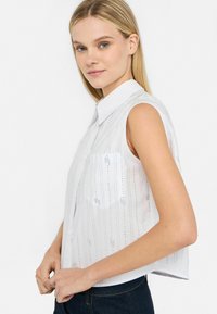 Blusa bianca senza maniche con colletto a bottoni, caratterizzata da righe verticali testurizzate e una tasca sul petto, realizzata in tessuto leggero.