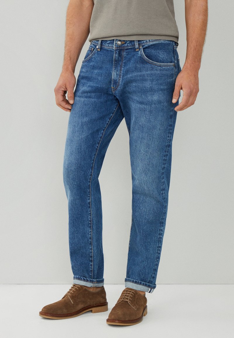Jeans de denim azul claro con corte recto, que presentan un diseño clásico de cinco bolsillos y dobladillos enrollados, combinados con zapatos de ante marrón.
