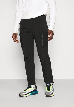 Mann trägt schwarze Cargohosen mit seitlichen Reißverschlusstaschen und neonakzentuierte Sportschuhe, steht vor einfachem Hintergrund.