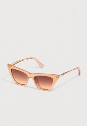 Lunettes de soleil en forme d’œil de chat avec des montures pêche translucides et des verres dégradés marron sur un fond clair.