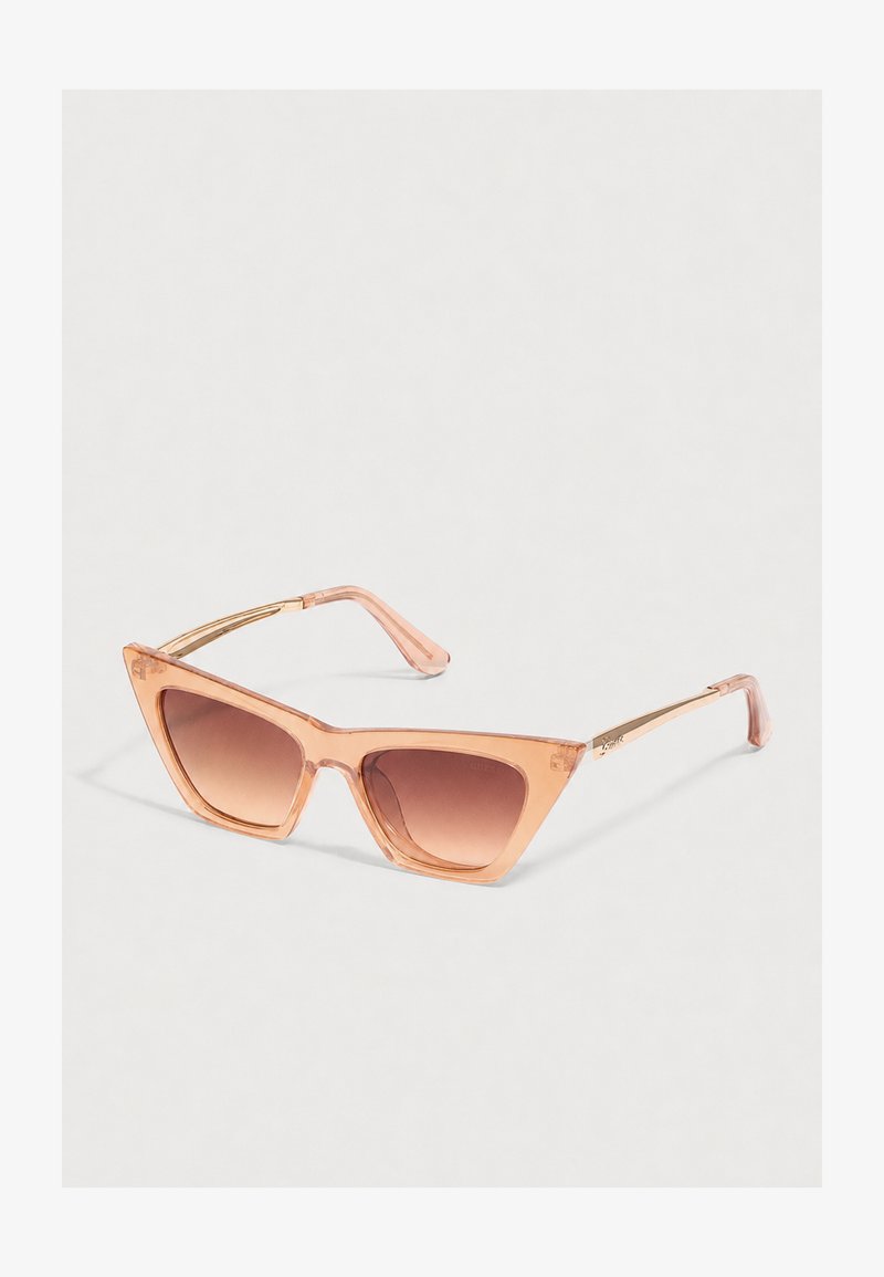 Lunettes de soleil en forme d’œil de chat avec des montures pêche translucides et des verres dégradés marron sur un fond clair.