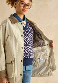 Jachetă bej cu guler din catifea, având nasturi tip snaps și căptușeală cu un model. Sub aceasta, un pulover albastru cu un design geometric roz.