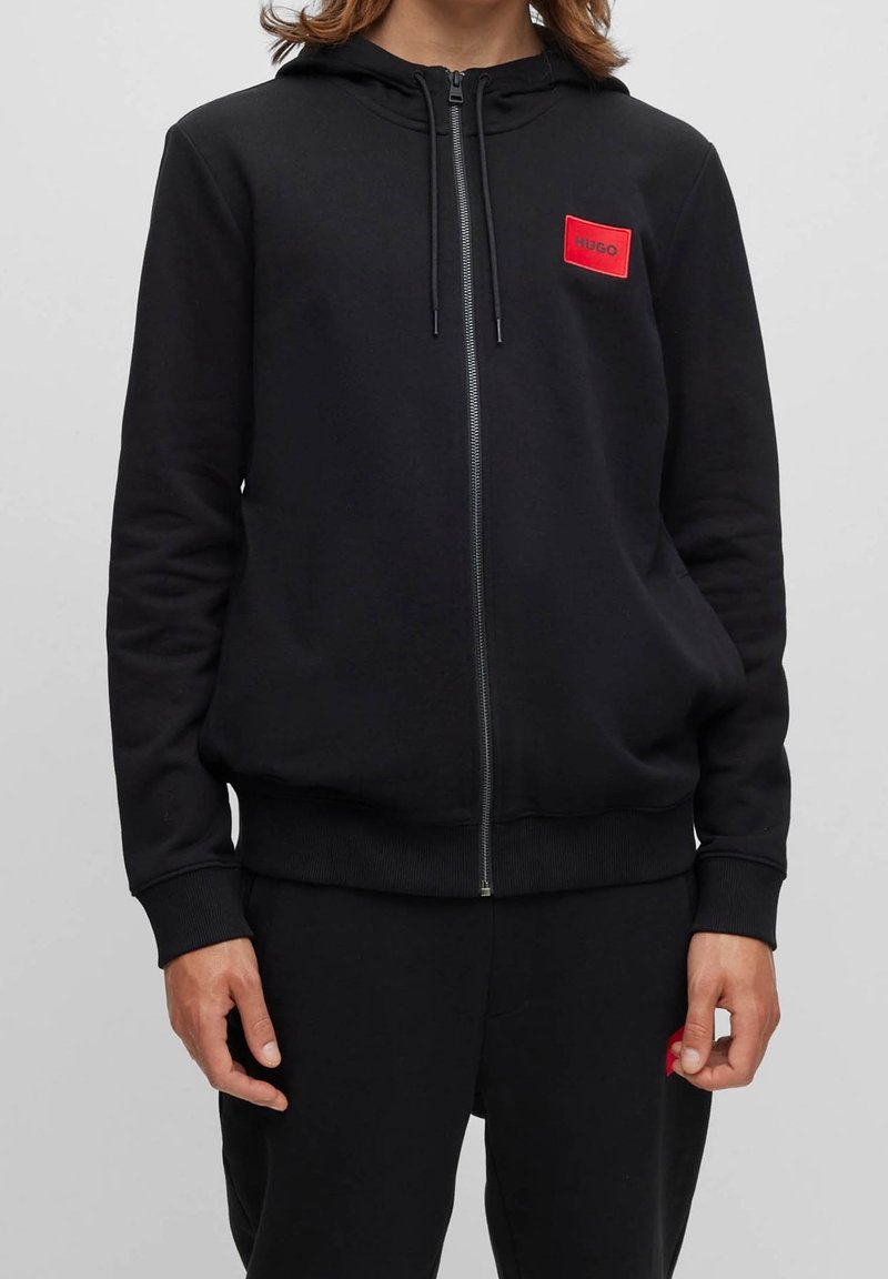 Personne portant un sweat à capuche noir zippé, avec un petit patch logo carré rouge sur la poitrine portant l'inscription "HUGO."