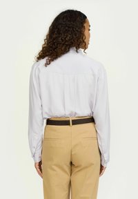 Lange mouwen lichtgrijs overhemd met een yoke-detail, gedragen met tan kleurige broek en een bruine gestructureerde riem, getoond vanuit de achteraanzicht.