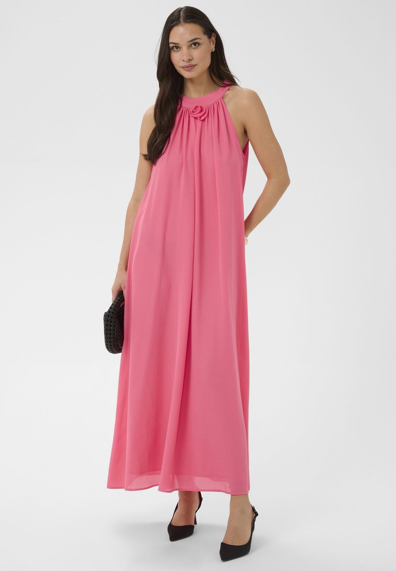 Femme portant une robe maxi rose sans manches avec un col froncé et un détail en forme de rose, tenant une pochette noire tissée, debout en talons noirs.