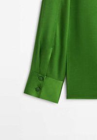Poignet de chemise vert à manches longues avec deux boutons verts assortis sur un tissu lisse, présenté sur un fond blanc.