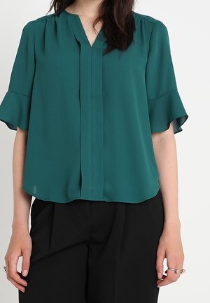 Femme portant un chemisier turquoise à manches courtes avec des manches à volants et un pantalon noir taille haute, debout, les bras détendus.